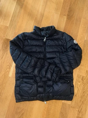Äkta Moncler jacka - Snygg svart dunjacka från Moncler med hög krage och två framfickor med dragkedja. Jackan har quiltad design och Moncler-logga på ärmen. Perfekt för kalla vinterdagar och riktigt lätt att bära tack vare det tunna dunet.perfekt för kvinnor under hösten och vintern. Så klar är den riktig och har kvittot som bevis, orginal pris 5495kr