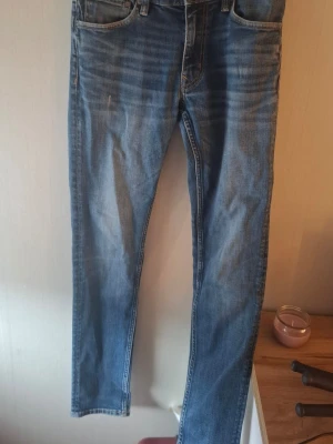 Blå raka jeans med slitningar - Säljer ett par klassiska blå jeans med raka ben och diskreta slitningar framtill. Jeansen har fem fickor och tydliga sömmar, samt en ljus tvätt som ger en avslappnad look. Perfekta till vardags och passar till det mesta i garderoben.