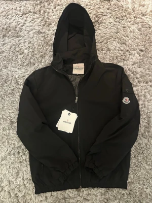 Svart Moncler vindjacka  - Snygg svart vindjacka från Moncler med luva och dragkedja framtill. Jackan har Moncler-logga på ärmen. Jackan är helt ny i storlek M, perfekt för hösten 🍁🍂