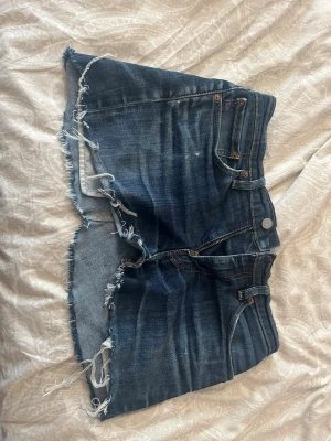 Mörkblå jeanskjol med fransig kant - Säljer ett par mörkblå jeanskjol med rå, fransig kant och klassisk femficksdesign. Kjolen har dragkedja och knapp framtill samt detaljerade sömmar och en liten etikett bak. Perfekta för sommaren! Obs hade ej lapp inuti men skulle gissa storlek S.