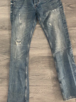 Blå slitna jeans med raka ben - Säljer ett par blå jeans med raka ben och slitningar på både lår och knä. Jeansen har en klassisk femficksmodell och tydliga tvättade detaljer för en trendig, avslappnad look. Perfekta för dig som gillar en casual streetstil. 31 midjan och 32 längd 