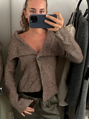 Brun stickad kofta med knapp - Säljer en brun stickad kofta med en stor dekorativ knapp framtill. Koftan har en bred krage som kan bäras off-shoulder och långa ärmar med avslappnad passform. Perfekt för lager på lager och en chill look.