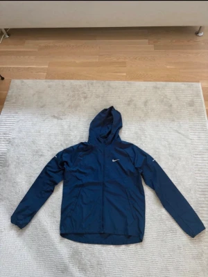 Blå Nike vindjacka med huva - Snygg blå vindjacka från Nike med huva och dragkedja framtill. Jackan har två sidofickor och en vit Nike-logga på bröstet. Perfekt för blåsiga dagar och enkel att matcha med andra plagg. Lätt och smidig design.