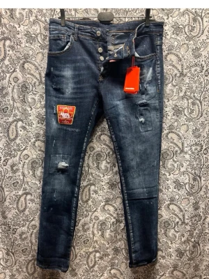 Mörkblå slitna jeans med patch - Säljer ett par mörkblå jeans med slitningar och en röd patch på vänster lår. Jeansen har skinny fit, knappar i gylfen och detaljerade slitningar på både fram- och baksida. Snygg tvätt och trendig look, perfekta för dig som gillar streetstyle.