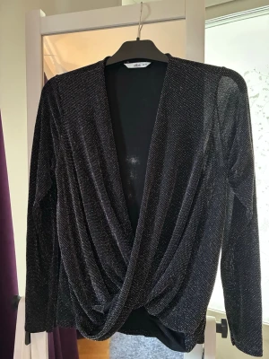 Glittrig omlottopp i svart - Snygg svart omlottopp med glittrigt tyg och lång ärm. Toppen har en djup v-ringning och ett draperat omlottparti framtill som ger en cool och trendig look. Perfekt för dig som vill sticka ut lite extra.