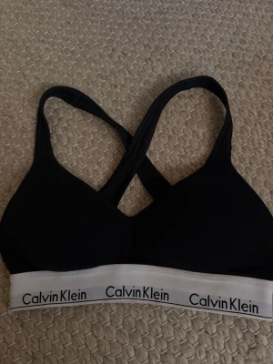 Svart Calvin Klein Bh - Svart pusch up bh från Calvin Klein med vadderade kupor och korsade axelband i ryggen. Mjuk och stretchig modell som passar perfekt till träning eller chill.