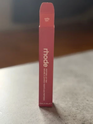 rhode Peptide Lip Tint Salty Tan - Säljer ett rhode Peptide Lip Tint i färgen Salty Tan. Förpackningen är avlång och rosa med vit text. Innehåller 10 ml. Perfekt för dig som vill ha återfuktade läppar med en hint av färg.
