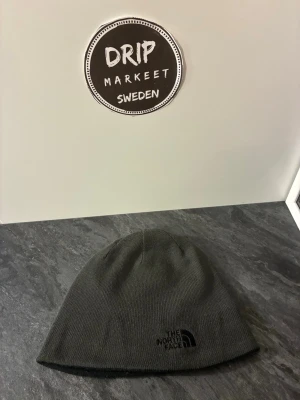 Mörkgrå mössa från The North Face - Snygg och enkel mörkgrå mössa från The North Face med diskret logga broderad framtill. Tillverkad i mjukt och stretchigt material som sitter skönt på huvudet. Perfekt för kalla dagar och passar till de flesta outfits.