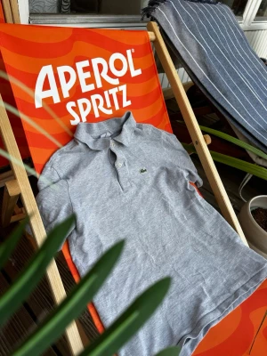 Grå pikétröja från Lacoste - Klassisk grå pikétröja från Lacoste med krage och två knappar framtill. Tröjan har korta ärmar och den ikoniska gröna krokodilloggan på bröstet. Perfekt till sommaren för en clean och stilren look.
