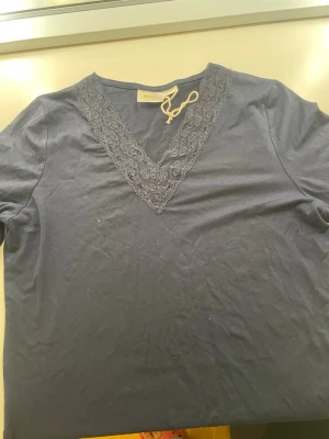 Mörkblå topp med spets från Gina Tricot - Snygg mörkblå topp från Gina Tricot med v-ringning dekorerad med spetsdetaljer. Tillverkad i mjuk bomullsmix och har korta ärmar. Perfekt för dig som gillar en enkel men ändå elegant stil.