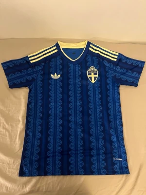 Blå Sverige fotbollströja Adidas - Säljer en blå fotbollströja från Adidas med Sveriges landslagsemblem på bröstet. Tröjan har gula detaljer vid krage och axlar samt ett unikt mönster i ljusblått. Tillverkad i lätt och ventilerande material, perfekt för träning eller match.