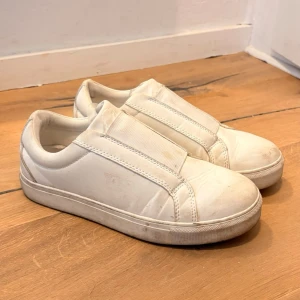 Vita slip-on sneakers utan snörning - Stilrena vita slip-on sneakers med elastiska band istället för snören. Skorna har en låg profil och en tjock vit sula. Tillverkade i syntetmaterial med enkel och clean design som passar till det mesta.