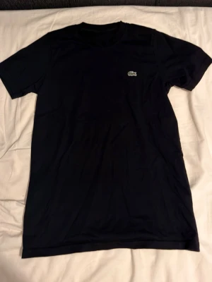 Svart t-shirt från Lacoste - Snygg svart t-shirt från Lacoste med den klassiska krokodilloggan på bröstet. T-shirten har rund halsringning och korta ärmar. Perfekt basplagg till garderoben och enkel att matcha med allt.