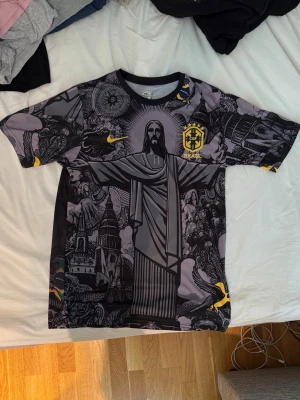 Brasil Neymar Jr. Fotbollströja  - Snygg svart Brasilien matchtröja från Nike med Neymar Jr. och nummer 10 på ryggen. Tröjan har ett unikt grafiskt tryck med Kristusstatyn och brasilianska motiv i grått och gult. Korta ärmar och rund hals. Perfekt för fotbollsfans! Sitter väldigt skin och tajt på kroppen. Efterliknar mer som storlek S