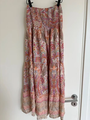 Mönstrad långkjol i viskos - Bohemisk långkjol i mjuk viskos med färgglatt paisley- och blommönster i rosa, orange, beige och lila toner. Kjolen har smock i midjan för skön passform och ett flowy fall som ger en avslappnad vibe.