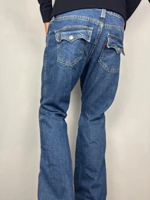 Levi’s Noughties Bootcut Jeans Blå W34 - Levi’s ”Noughties Bootcut” jeans med unika lockfickor och tidlös 00-talslook. Färg: Blå mellantvätt Passform: Bootcut Storlek: W34 Skick: Mycket bra begagnat skick (A/B Grade). Mått [platt] (±1 cm): A: 47 cm B: 95 cm D: 31 cm C: 26 cm För korta på mig i bilden som är 189 cm, har fått dra ner jeansen rejält i syfte att visa passform. Tveka inte att höra av dig vid frågor eller funderingar. Skickas snabbt! 