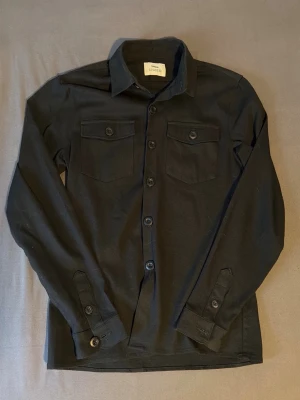 Ciszere Overshirt - Stilren svart overshirt med knäppning framtill och två bröstfickor med lock och knapp. Klassisk krage och långa ärmar med manschetter. Perfekt att bära över en t-shirt för en avslappnad look. Modellen är 180cm och väger ca 65kg