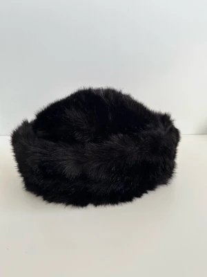 Wigéns - svart mössa i fuskpäls - storlek M / 57 - Stilren svart mössa i kvalitativ fuskpäls från anrika svenska pälstillverkaren Wigéns i storlek 57 / M (inre omkrets 57cm). Klassisk design med rundad topp och brett band runtom. Supermjuk och len fuskpäls. En tidlös klassiker som kommer förhöja outfiten under alla kallare dagar. Mycket gott skick, som ny!