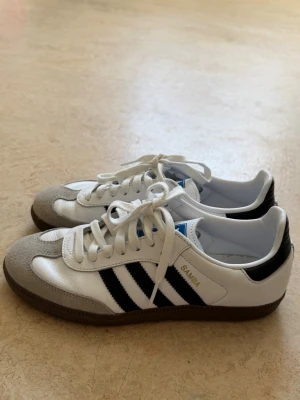 Adidas Samba sneakers - Klassiska Adidas Samba sneakers i vitt läder med svarta ränder och grå mockadetaljer på tån. Skorna har bruna gummisulor och vita skosnören. Perfekt för dig som gillar retrostil och sportig vibe. Sparsamt använda och i väldigt bra skick enligt mig🤍🙌🏼De är i storlek 39 1/3 men jag bär egentligen 38 i andra skor så de är ”mindre” i storleken.