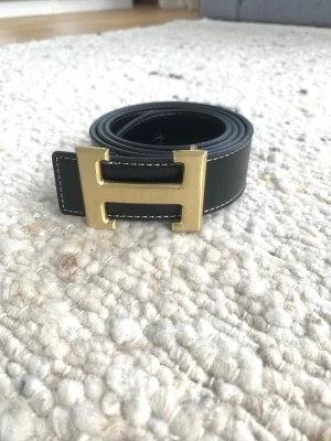 Svart skärp med H-guldspänne - Stilrent svart skärp med klassiskt H-format spänne i guldmetall. Skärpet har vita kontrastsömmar längs kanterna och är tillverkat i skinn. Perfekt designer accessoar för att lyfta din outfit.