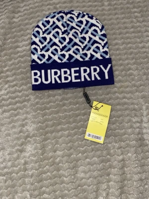 Blåvit Burbǝrry mössa  - Snygg blåvit mössa från Burbǝrry  med stort logotyp-tryck framtill och ett grafiskt mönster i olika blå nyanser. Tillverkad i mjukt stickat material som håller dig varm och ger en trendig look. Perfekt accessoar för dig som vill sticka ut.