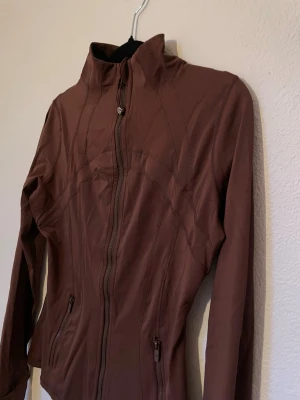  Lululemon st 6  - Snygg brun jacka från Lululemon med figurnära passform och dragkedja framtill. Jackan har långa ärmar, hög krage och två fickor med dragkedja. Perfekt för en aktiv livsstil och enkel att matcha med olika outfits.