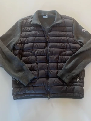 Mörkgrå cardigan - Snygg mörkgrå moncler cardigan med quiltad front och rygg samt stickade ärmar och krage. Jackan har dragkedja framtill, två sidofickor med dragkedja och en liten logga på vänster ärm. Perfekt för kalla vinterdagar och ger en modern look. Passar runt 170-180 cm