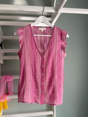 Rosa ärmlös blus med knappar - Säljer en söt rosa blus med vita mönster och knappar framtill. Blusen är ärmlös med volangdetaljer vid axlarna och har en djup v-ringning. Materialet känns lätt och luftigt, perfekt för varma dagar. Passar dig som gillar boho-vibbar och vill ha något färgglatt. Finns några lösa trådar (se sista bild) men annars inga defekter.