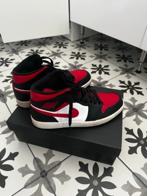 Svarta och röda Nike Air Jordan 1 - Säljer ett par Nike Air Jordan 1 sneakers i svart, rött och vitt. Klassisk high-top modell med svart snörning, röd swoosh och detaljer samt vit mellansula. Tillverkade i läder med ikonisk vinge-logga på sidan. Perfekta för dig som gillar streetwear och sneakerskultur.