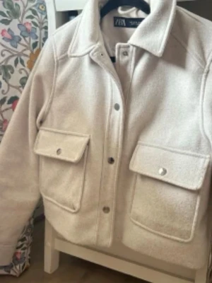 Vitgrå kappa Zara -  LITEN I STORLEK !!! Stilren beige overshirt från Zara i mjukt fleece-liknande material. Jackan har stora bröstfickor med lock och tryckknappar, klassisk krage och raka ärmar. Perfekt för lager på lager och enkel att matcha med olika outfits.