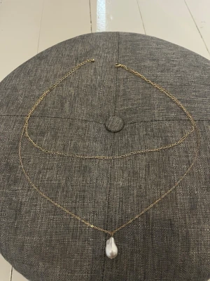 Guldhalsband med droppformad pärla - Snyggt halsband med tunn guldfärgad kedja och en stor droppformad vit pärla som hänge. Kedjan har en enkel och stilren design som passar till många olika looks. Perfekt accessoar för dig som gillar klassiska smycken med en modern twist.