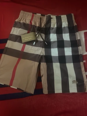 Beige-rutiga shorts med snörning XL - Säljer ett par beige shorts med klassiskt rutigt mönster i svart, vitt och rött. Shortsen har snörning i midjan och elastisk resår för skön passform. Perfekta för sommaren och har en modern, avslappnad stil.