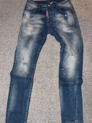 Blå dsquard2 jeans med färgstänk och slitningar - Säljer ett par blå dsquard2 jeans med coola slitningar och färgstänk i vitt och rött. Jeansen har en avslappnad passform och klassisk femficksdesign. Perfekta för dig som gillar en trendig och unik streetstil.