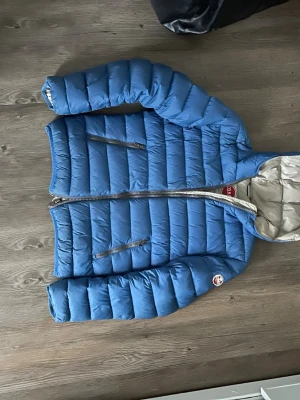 Blå dunjacka från Colmar Originals - Snygg blå dunjacka från Colmar Originals med quiltad design och två dragkedjeförsedda fickor framtill. Jackan har en huva och insidan är ljusgrå. Perfekt för kalla vinterdagar och riktigt skön att bära.