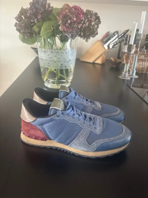 Valentino rockrunner - Snygga sneakers i blått tyg och mocka med röda och beige detaljer på hälen. Skorna har en beige sula med svarta inslag och blå skosnören. Perfekta för dig som vill ha en unik och trendig look.