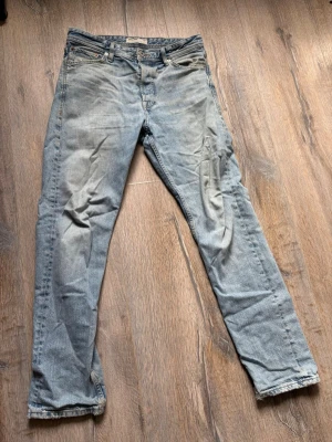 Ljusblå  jeans med fransig kant - Säljer ett par ljusblå jeans med rak passform och fransig kant vid bensluten. Jeansen har klassisk femficksdesign, knappgylf och är tillverkade i denim. Perfekta för en avslappnad och trendig look.