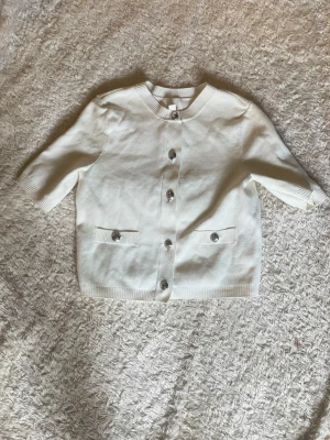 Vit kofta från H&M, XS - Stilren vit kofta från H&M i storlek XS. Koftan har rund halsringning, korta ärmar och silverfärgade knappar framtill. Två små fickor framtill med dekorativa knappar. Perfekt att slänga över en topp för en clean look.