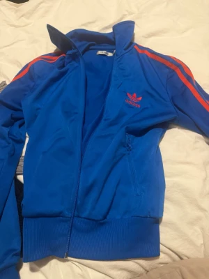 Blå Adidas track jacket med röda ränder - Snygg äldre modell blå track jacket från Adidas med röda ränder längs ärmarna och klassisk Adidas-logga på bröstet. Jackan har dragkedja framtill, hög krage och ribbade muddar. Perfekt för en sportig och retro look.