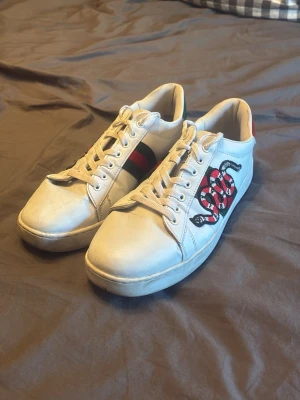 Vita sneakers med orm och ränder - Snygga vita sneakers med broderad röd och svart orm på sidan samt klassiska gröna och röda ränder. Skorna har vita snören och en låg profil som passar till många olika stilar. Materialet är slätt läder och sulan är vit. OBS!! Hål i hällen
