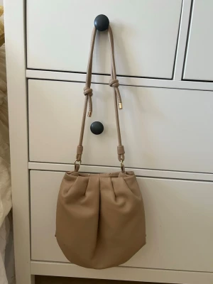 Beige bucket bag med knytband - Säljer en beige bucket bag med mjuk form och justerbara knytband som axelrem. Väskan har guldfärgade detaljer och är gjord i ett slätt syntetmaterial. Perfekt för dig som gillar minimalistisk och stilren design. Finns lite mindre svarta fläckar som kan ses på bilderna.