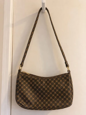 Brun axelremsväska med monogrammönster - Snygg brun handväska med klassiskt monogrammönster i guldtoner. Väskan har en avlång, mjukt rundad form och justerbar axelrem. Stängs med dragkedja och har en rymlig insida. Perfekt för dig som gillar tidlös och trendig design.