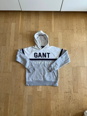 Gråvit hoodie från GANT med huva - Snygg hoodie från GANT i grått och vitt med mörkblå detaljer och stor logga på bröstet. Klassisk huva och känguruficka framtill. Mjuk bomullsblandning som känns skön mot huden. Perfekt för en avslappnad och trendig look.