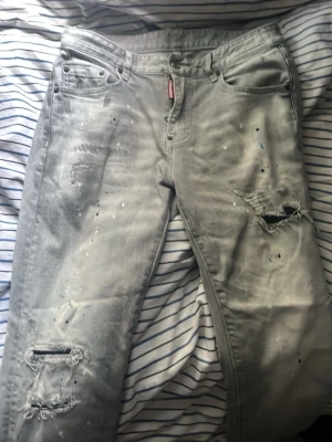 Grå slitna jeans med hål och stänk från dsqueard - Säljer ett par grå jeans med slitningar, hål och färgstänk för en riktigt edgy look. Jeansen har klassisk femficksmodell och raka ben. Perfekta för dig som gillar streetwear och vill sticka ut med en unik stil.