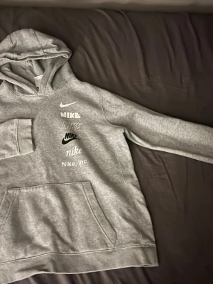 Grå hoodie från Nike med loggor - Snygg grå hoodie från Nike med flera olika Nike-loggor tryckta på bröstet. Hoodien har en klassisk känguruficka framtill och mjukt, borstat innerfoder. Perfekt för en avslappnad och sportig stil. 158-170