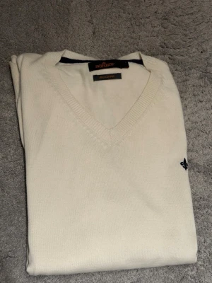Vit stickad tröja från Morris - Klassisk vit stickad tröja från Morris Stockholm i mjuk pima cotton. Tröjan har V-ringning och en liten mörkblå logga broderad på bröstet. Perfekt för dig som gillar stilrena och tidlösa plagg.