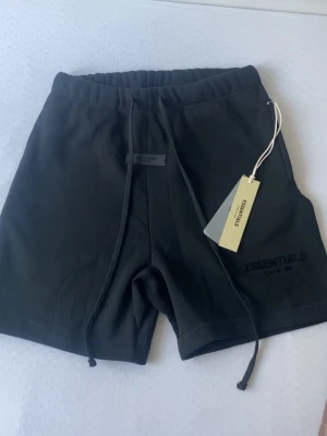 Svarta shorts från Essentials - Svarta shorts från Essentials Fear of God med elastisk midja och snörning. Klassisk och enkel design med logga på vänster ben. Perfekta för en avslappnad och trendig stil.