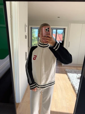 Vit och svart ziptröja med ränder - Säljer en sportig ziptröja i vitt och svart med högt krage och svarta ärmar. Tröjan har vita och svarta ränder på ärmarna samt en röd S på bröstet. Ribbstickade muddar vid ärmslut och nederkant. Perfekt till en avslappnad look.