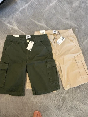 Två par cargoshorts i grönt och beige - Säljer två par cargoshorts från 157 i färgerna mörkgrön och beige. Båda har flera praktiska fickor på sidorna, bälteshällor och klassisk cargomodell. Perfekta för en avslappnad och trendig stil. Materialet är slitstark bomull.