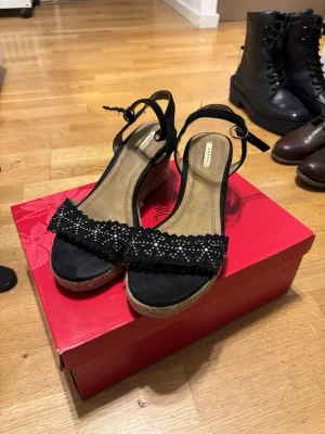 Svarta kilklackssandaler med nitar  - Snygga svarta sandaler med kilklack och öppen tå. Remmarna är dekorerade med små silverfärgade nitar och har ett vågigt mönster. Sandalerna har justerbar ankelrem och sulan är i naturfärgat material som liknar jute. vid frågor eller om ni vill ha fler bilder är det bara att höra av sig!