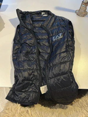 Svart dunväst från EA7 Emporio Armani - Snygg svart dunväst från EA7 Emporio Armani med quiltad design och diskret logga på bröstet. Västen har dragkedja framtill och är lätt vadderad för extra värme. Perfekt att slänga över en hoodie eller tröja för en sportig look.